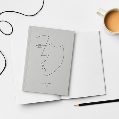 A5 notebook with grey softcover, from the brand Kinshipped. The cover has a minimalist drawing of a face and the words 'Hello You' written with gold foiled. 